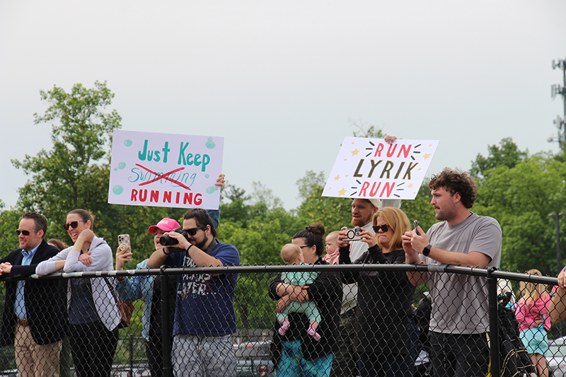A group of parents stand behind a chain link fence holding up signs to cheer on their favorite runners.