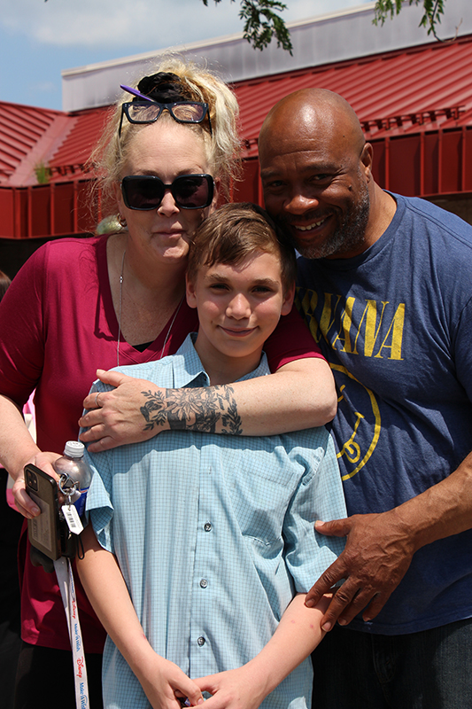 A fifth-grade boy smiles as two adults hug him.