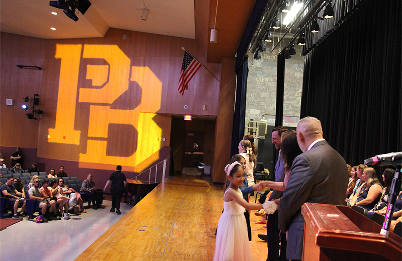 With a large PB reflected on a wall, fifth-grade students walk across a stage and shake hands with adults.