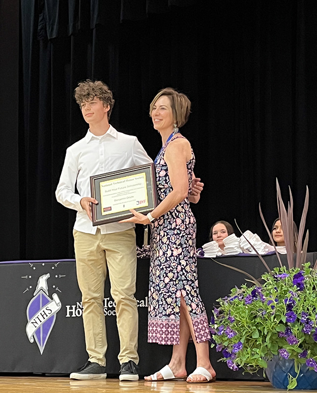A high school senior with short dark hair, wearing a button down white shirt and khaki pants receives a plaque from a woman in a long printed dress. They are on a stage and smiling.
