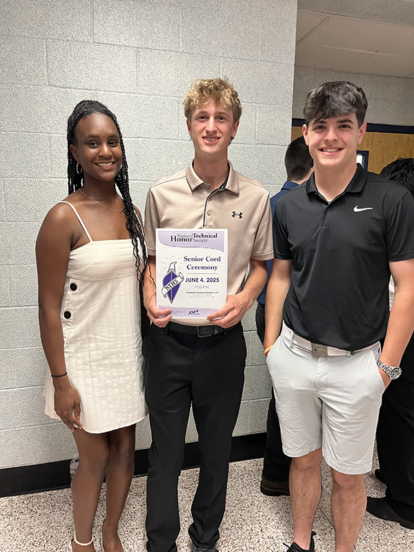 Three high school students stand and smile. The young man on the right is wearing a black polo shirt and shite shorts. The young man in the center is wearing adress sirt an dblack pants. He has blonde hair and is holding a program. The young woman on the left is wearing a white dress, has long dark hair and is smiling.