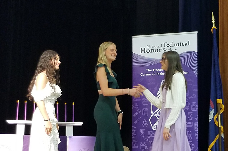 A young woman wearing a printed dress and white jacket shakes hands with a young woman on a stage. Another woman stands behind them. Everyone is smiling.