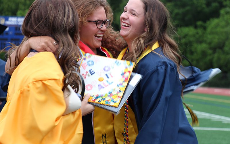 Three high school graduates in gold and blue caps and gowns hug each other and smile.