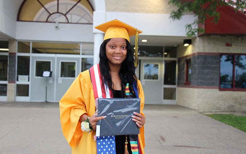 A young woman wearing a gold cap and gown smiles in front of a school building.