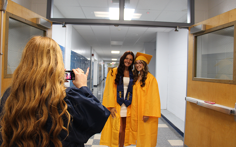 A young woman wit long blonde hair takes a photo of two young women wearing gold caps and gowns.