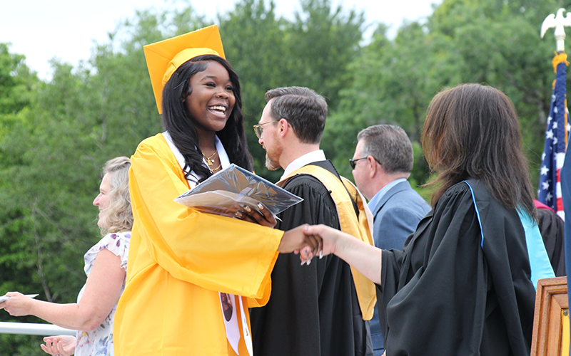 A young woman in a gold cap and gown shakes hands with a woman in a black gown.