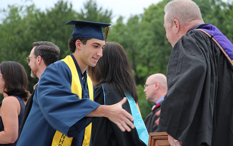 A young man in a blue cap and gown goes to shake the hand of a man in a black gown.