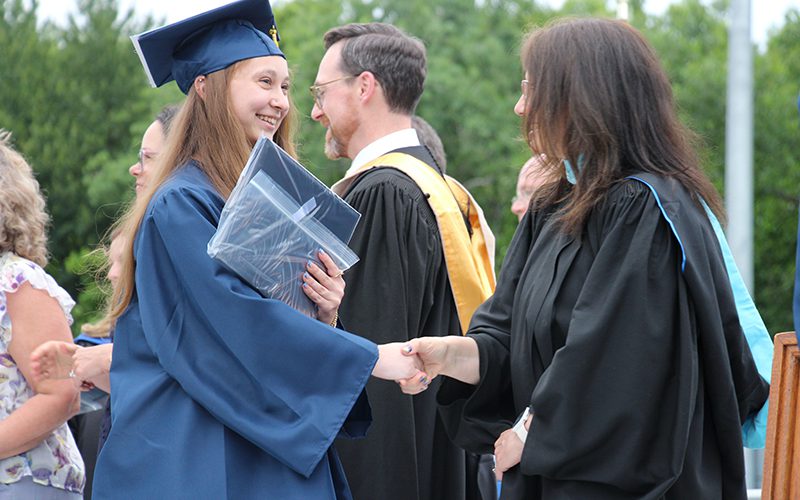 A young woman in a blue cap and gown shakes hands with a woman in a black gown.