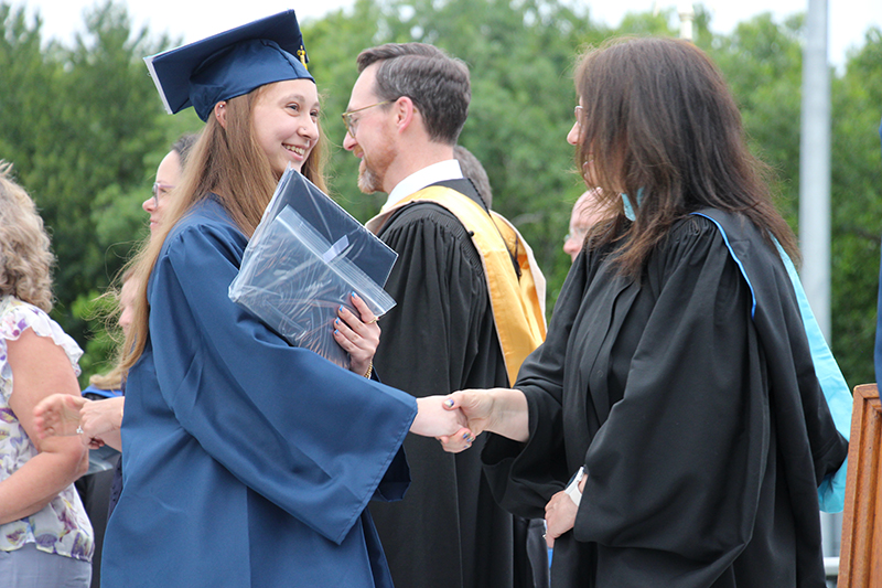 A young woman in a blue cap and gown shakes hands with a woman in a black gown.