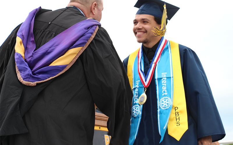 A young man in a blue cap and gown shakes hands with a man in a black gown.