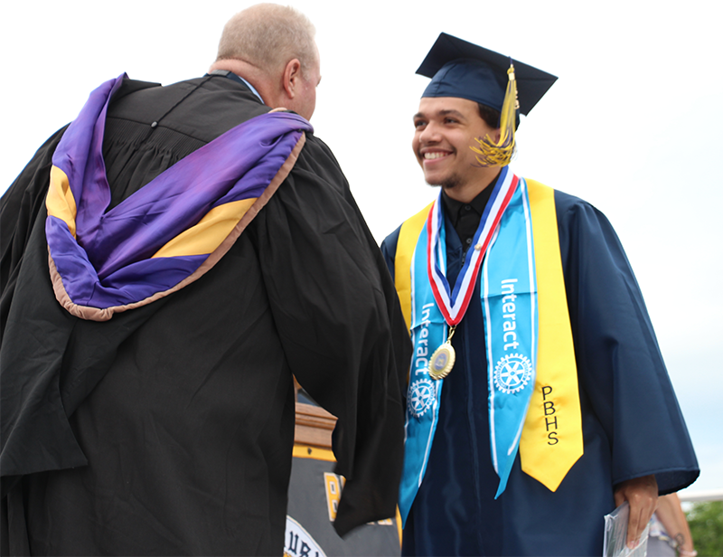 A young man in a blue cap and gown shakes hands with a man in a black gown.