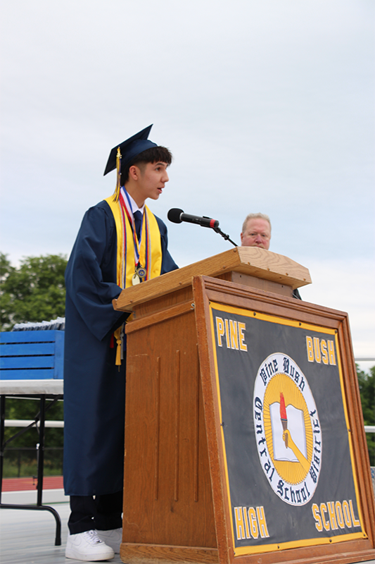 A young man in a navy blue cap and gown with gold stole talks at a podium.