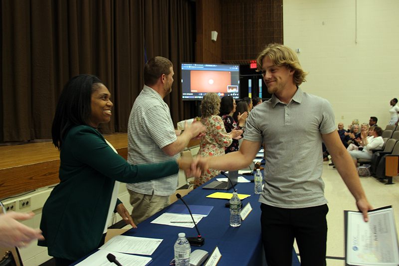 A young man smiles as he shakes the hand of a woman across the table. Other adults are clapping.