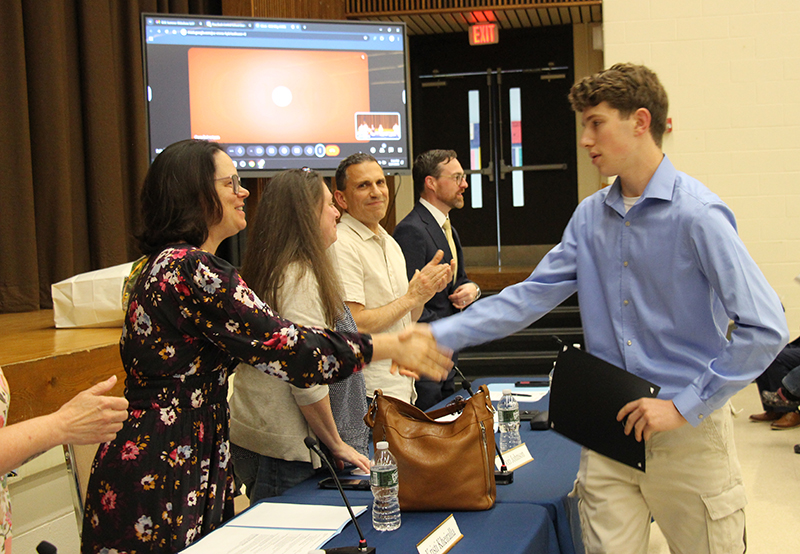 A young man shakes the hand of a woman as he walks down past other adults who are clapping and smiling.