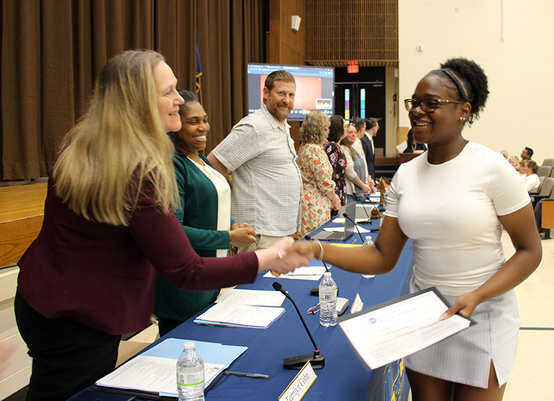 A young woman smiles as she shakes the hand of a woman across a table. There are other adults looking on on from down the other end of the table.
