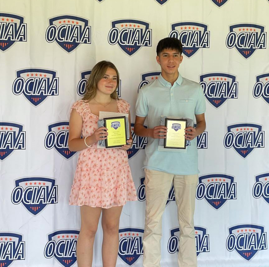 Two high school seniors - a young woman on the left and a young man on the right, stand in front of a backdrop that says OCIAA all around it. They are both smiling.