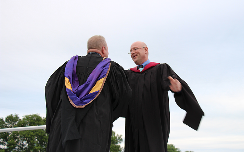 Two men in black graduation gowns embrace each other on a stage.