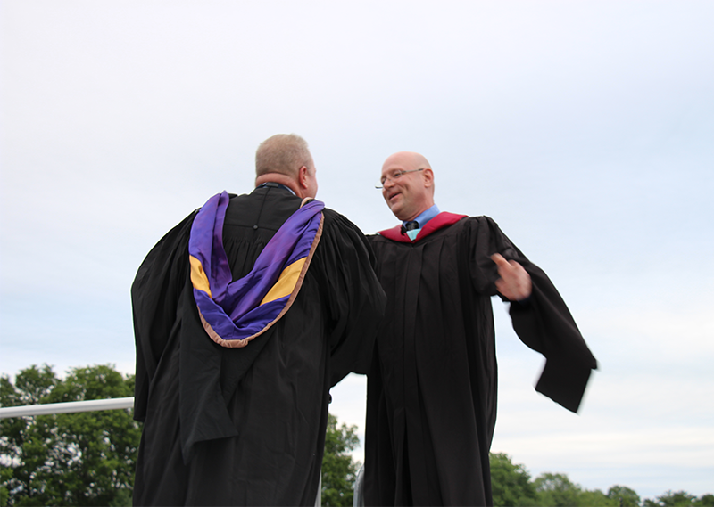 Two men in black graduation gowns embrace each other on a stage.