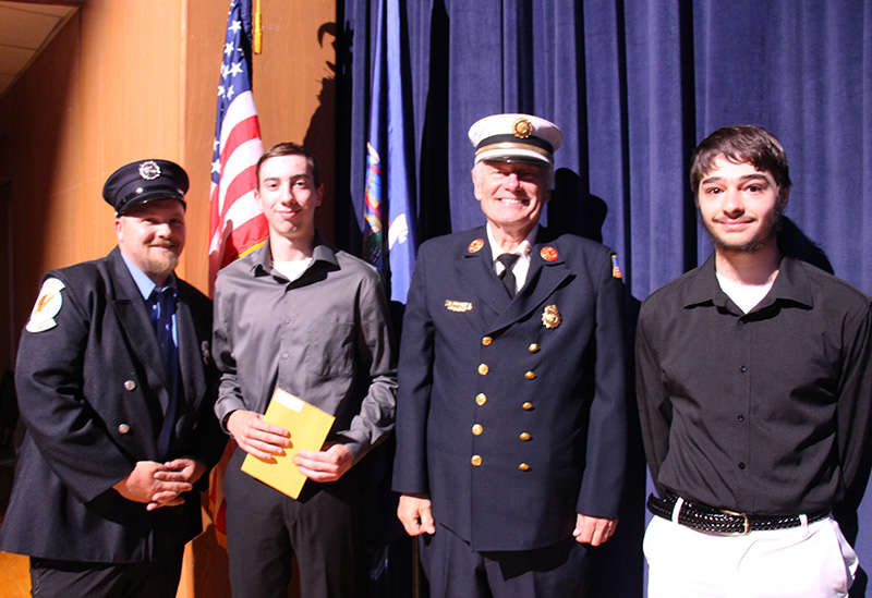Two men in formal firefighter uniforms stand next to two high school students.