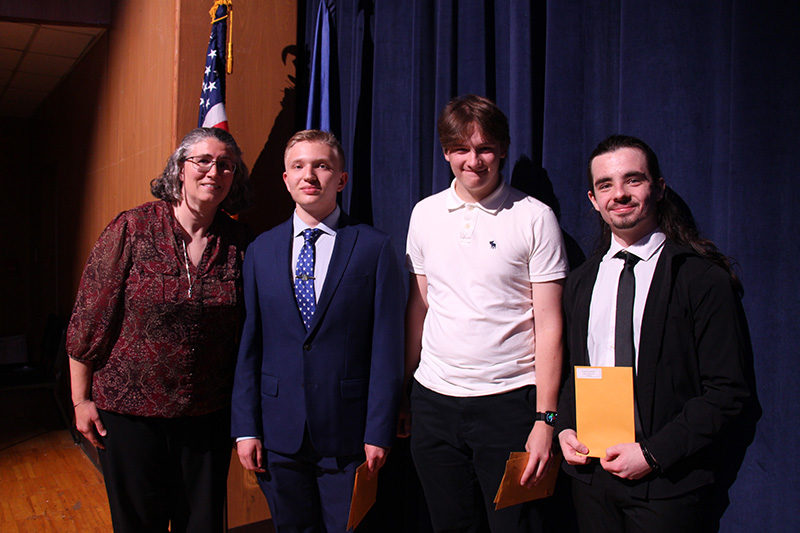 A woman stands on a stage with four high school seniors. They are all smiling.