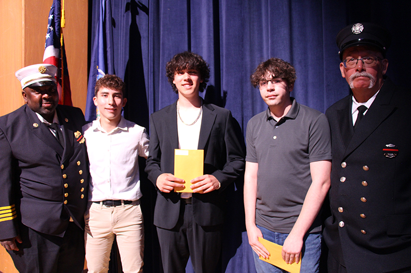 Three young men stand in the center with two men on either side, both wearing formal firefighter uniforms.