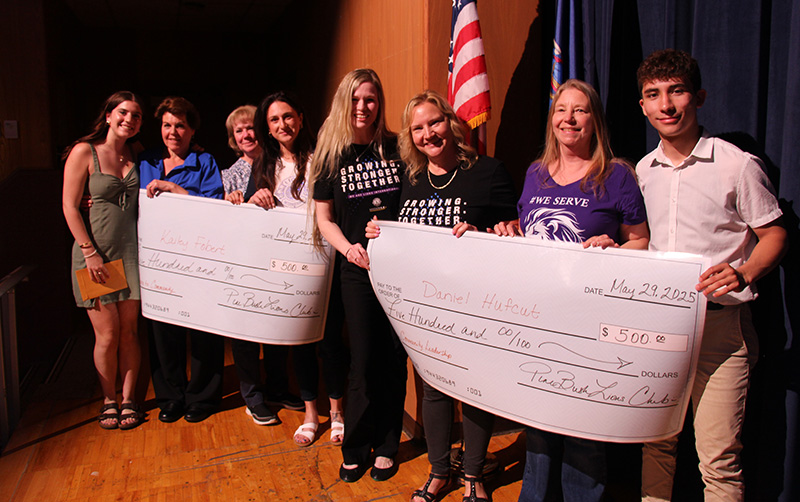 Six adults stand with two high school students. They have two oversized checks.