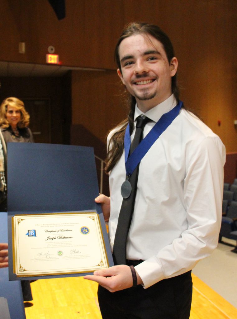 A young man smiles as he shows the certificate he just received. He is wearing a dress shirt and tie and he is smiling.