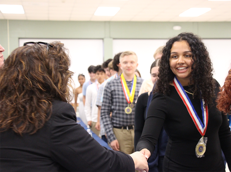 A young woman smiles as she shakes hands with a woman. There are other young people behind her.