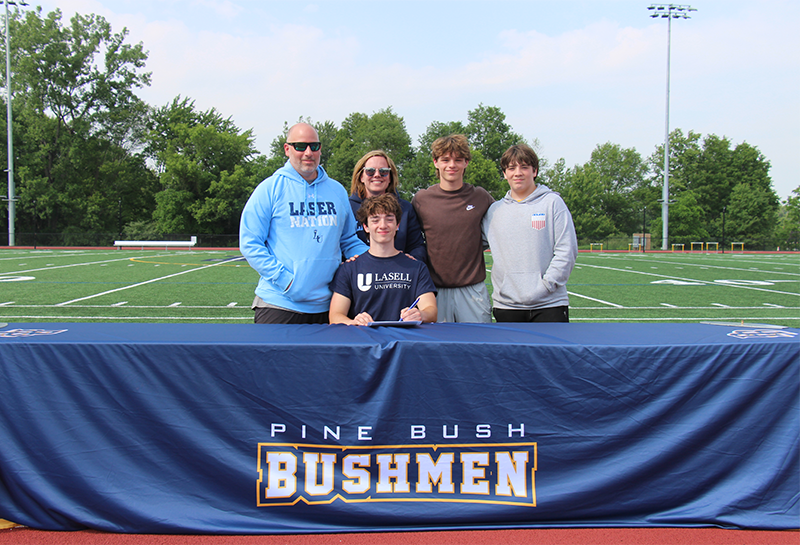 A young man smiles and sits at a table that says Pine Bush Bushmen. Behind him are a man, woman and two young men.