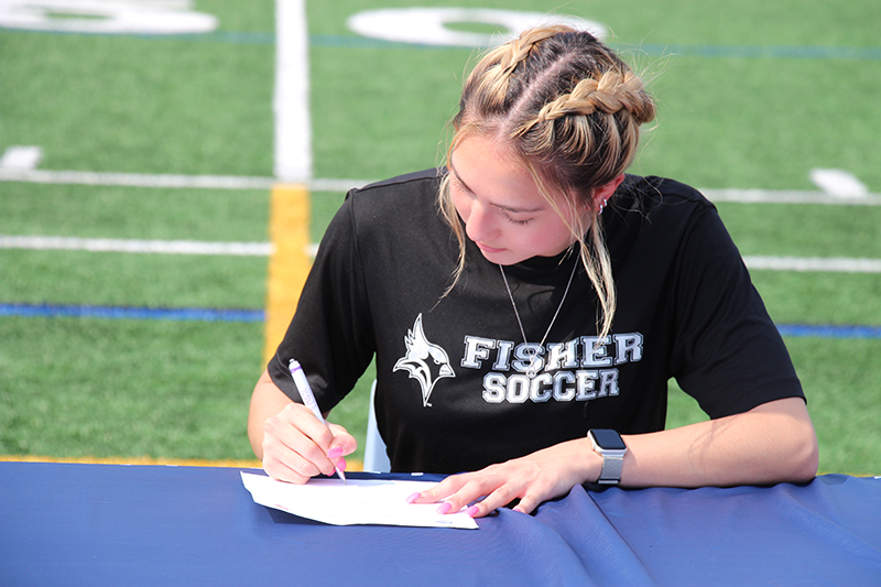 A young woman, with her hair in braids and wearing a black tshirt that says Fisher Soccer, signs a letter while siting at a table on an athletic field.