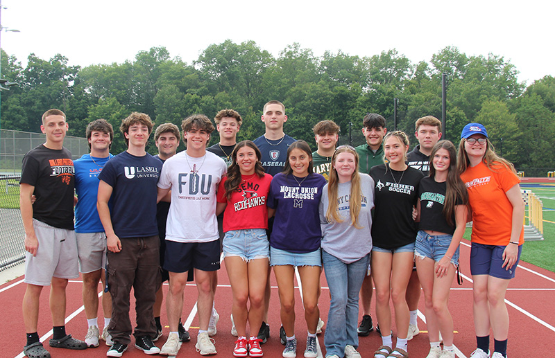 A group of about 15 high school seniors stand on a track, arm in arm, in two rows.