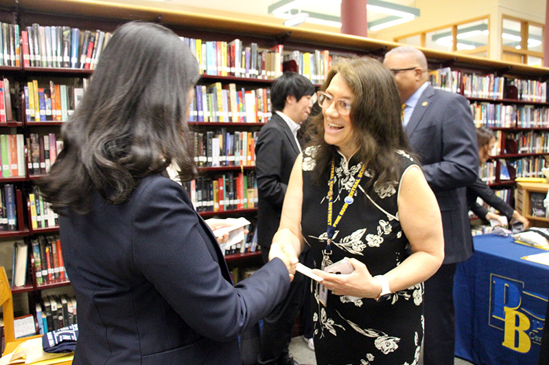 A woman with longer dark hair and glasses smiles and shakes hands with a woman in a dark blue suit.