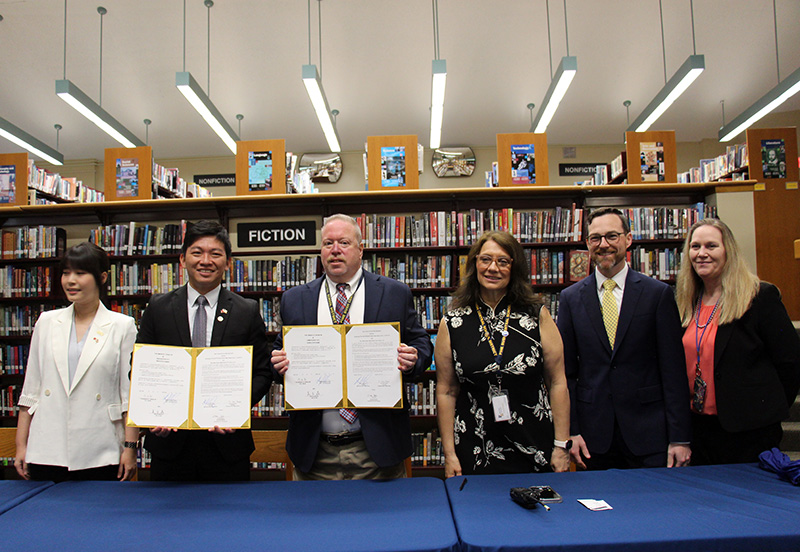 Six people stand behind a table. In the center, the two men hold up letters they've both signed. Everyone is smiling.