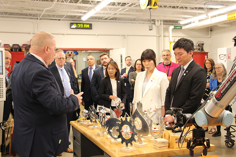 A man stands on the left and other adults stand around a table with many metal products made by students.