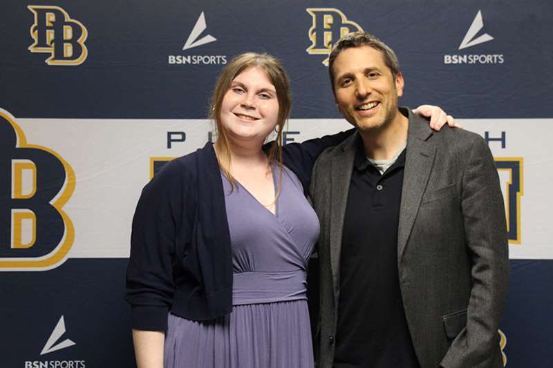A young woman with her light hair pulled back, wears a light purple dress and black sweater and stands with a man in a black shirt and sport jacket. They are smiling.
