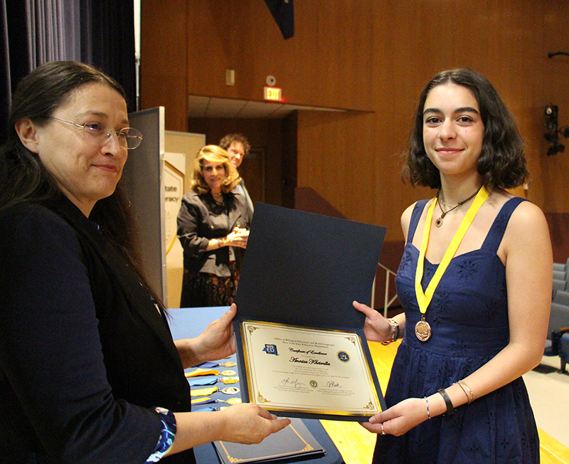A young woman in a blue dress wearing a gold medallion around her neck smiles and shows a certificate she is holding. A woman stands next to her smiling.