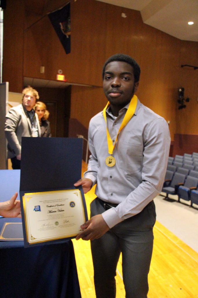 A young man dressed in a dress shirt holds a certificate he just received. He has a gold medallion around his neck.