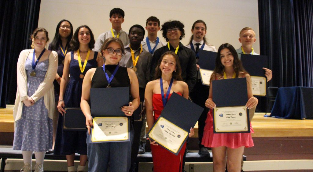 A group of 12 high school seniors stand on a stage, all holding  certificates they just earned. They are smiling.
