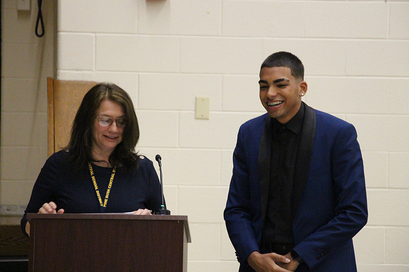A young man of high school age wearing all black smiles as a woman next to him stands at a podium and talks. 