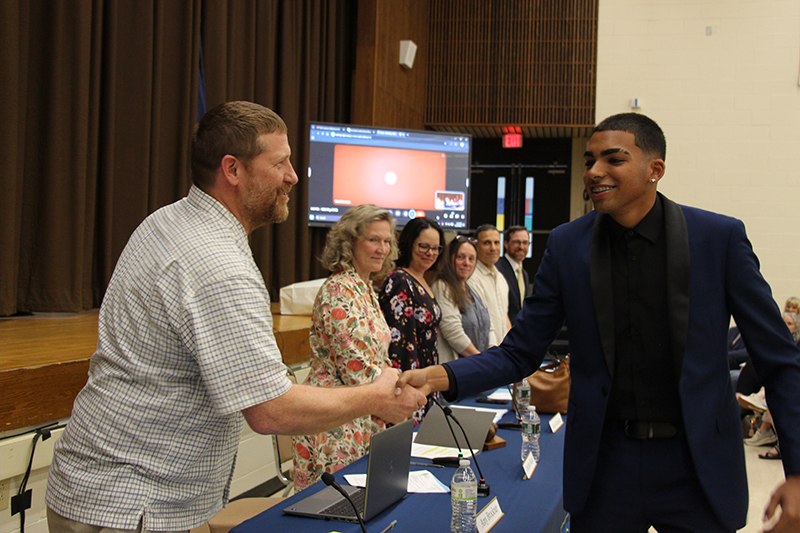 A young man dressed in black, shakes hands with a man behind a table. Other adults are looking on.