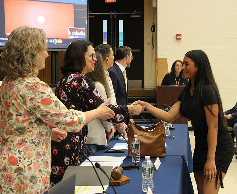 A young woman of high school age shakes hands with a woman behind a table. Others are looking on. She is dressed in a black dress and has long dark hair.