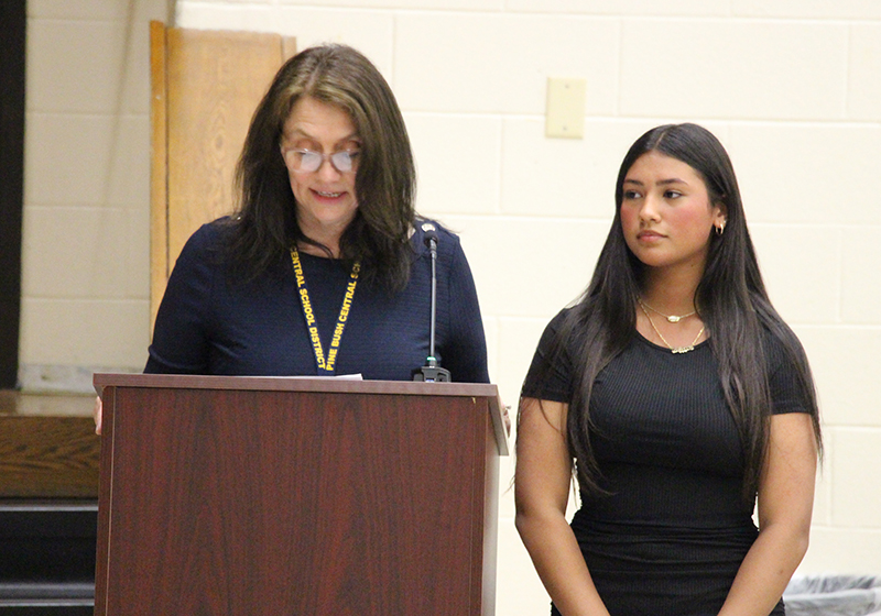 A young woman of high school age with long dark hair, wearing a black dress, stands next to a woman with mid-length hair who is speaking at a podium.