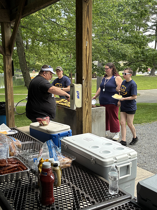 A man cooks on a large grill as high school kids wait in line for their food.