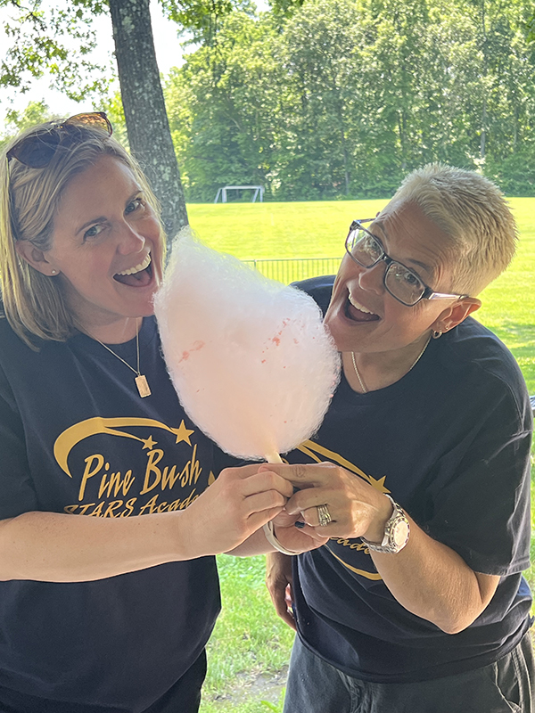 Two women smile as they hold one pink cotton candy and look like they are going to bite it.