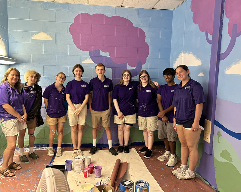 A woman on the left and seven high school students, all wearing khaki shorts and purple shirts, stand in front of a mural they created in a small elementary school room.