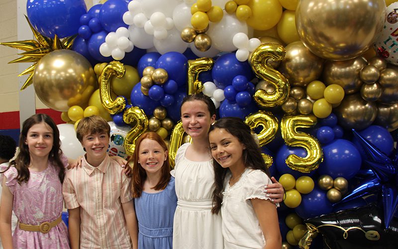 A group of five fifth-grade kids stand in front of a large blue and gold balloon display that says CES 2025. They are all smiling.