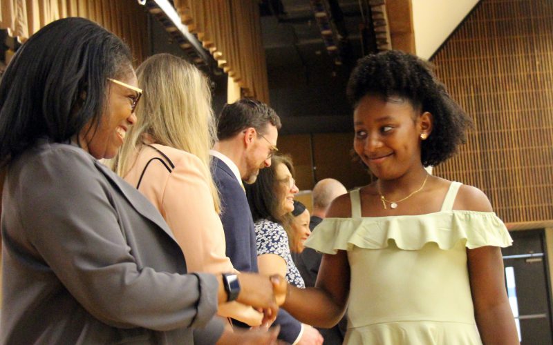 A fifth grade girl in a green dress smiles as she shakes hands with a woman.
