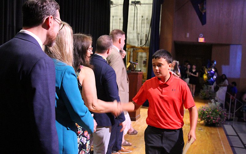 An eighth-grade boy in a red shirt shakes hands with a woman on a stage. Other adults are waiting to shake his hand.