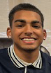 A young man with short dark hair smiles. He is wearing a blue shirt with a blue and white striped collar.