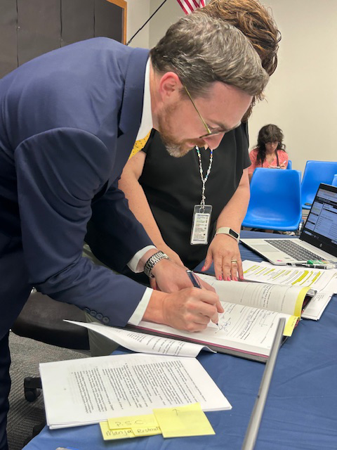 A man in a blue suit leans over a table and signs a book.